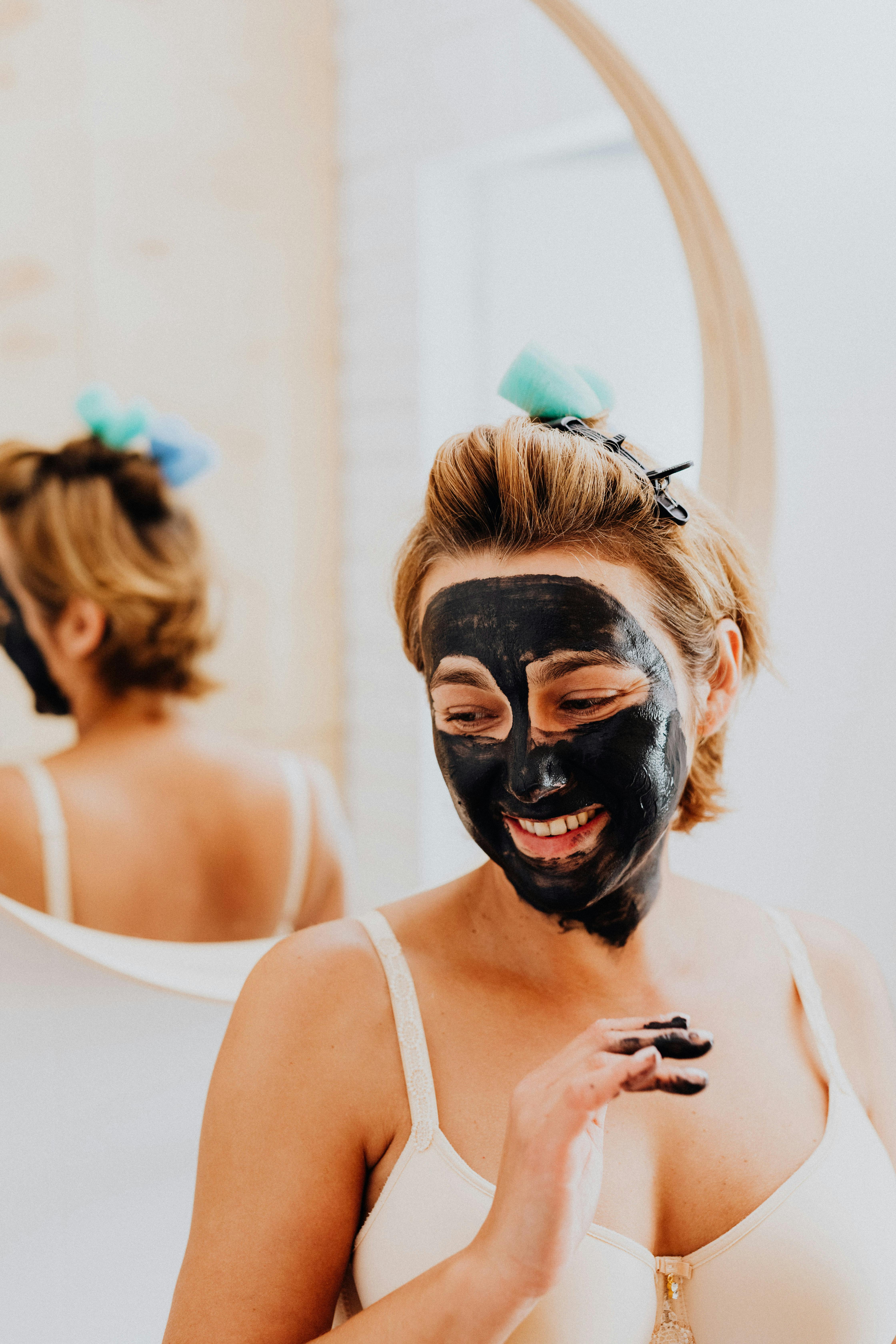 Woman applies black facial mask while smiling in a bathroom setting.