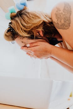 Woman with curlers washes face in a white sink, showcasing a daily skincare routine.
