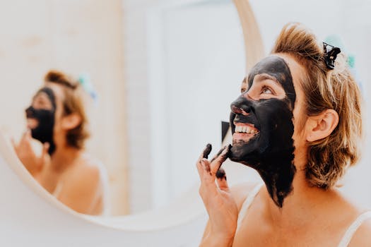 Happy woman applying black charcoal face mask in front of a mirror.