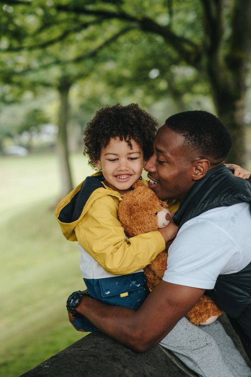 Free Tender African American male cuddling adorable little black boy with plush bear while sitting in nature with trees on blurred background Stock Photo