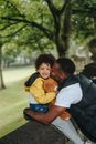 Side view of African American father hugging and kissing toddler boy carrying cute toy bear while sitting on concrete fence and smiling at camera