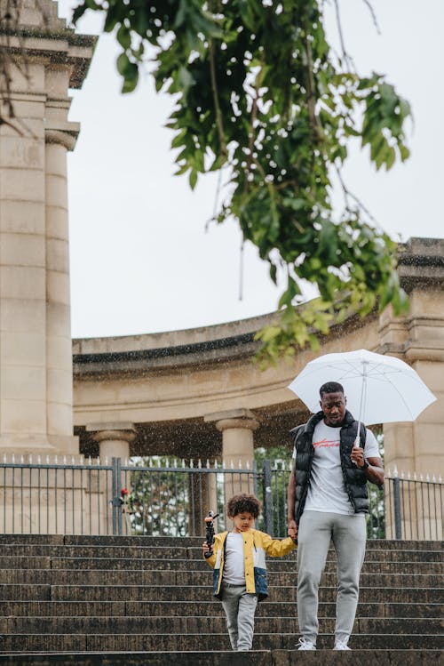 Young black man with umbrella walking downstairs with son