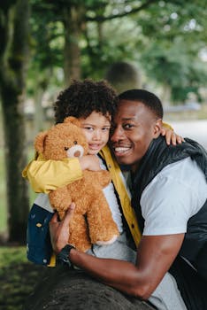 阳光明媚的户外公园里，一位快乐的父亲抱着儿子，手里还拿着泰迪熊