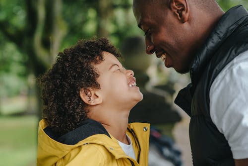 Side view of African American young father smiling while looking on joyful little son with closed eyes on blurred background
