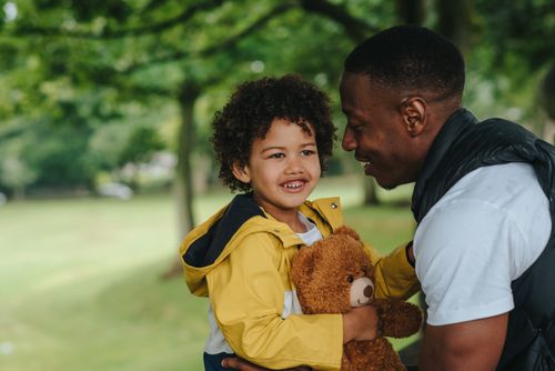 Free Happy little boy carrying toy bear and looking away while spending free time together with African American father in park Stock Photo