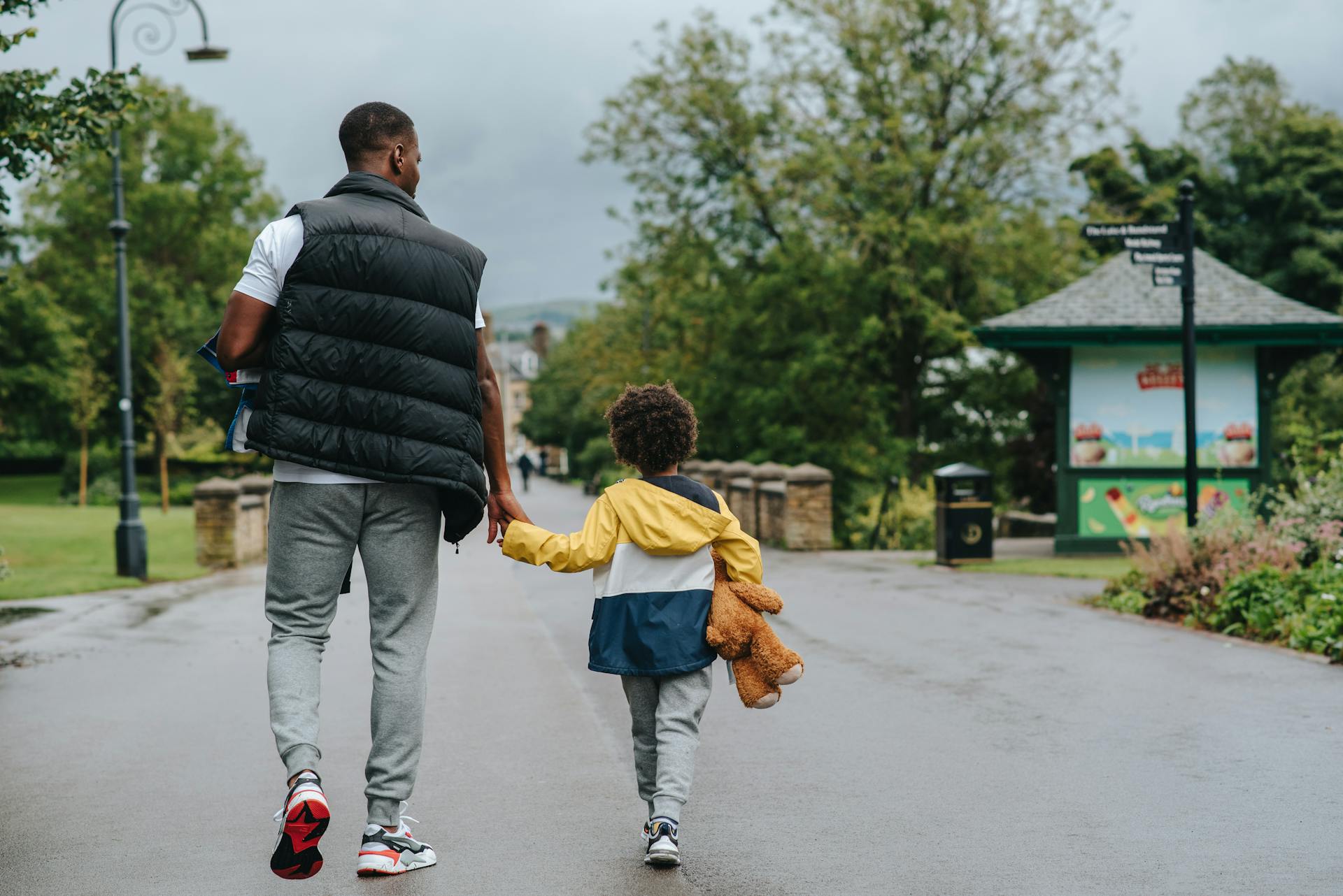 Back view of anonymous African American man holding child by hand while walking on urban roadway in daylight