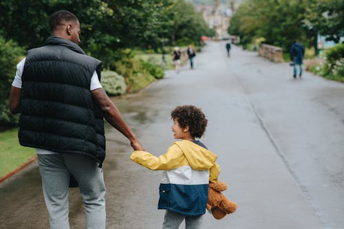 Back view of anonymous African American man holding cheerful son by hand while looking at each other on urban road