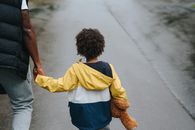 From above back view of crop anonymous ethnic father holding boy with toy bear by hand on asphalt roadway