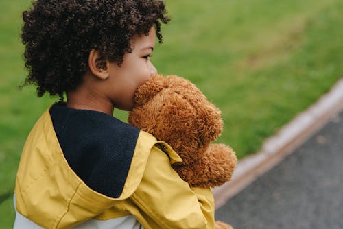 Back view of crop dreamy ethnic kid with toy bear looking away on walkway in summer