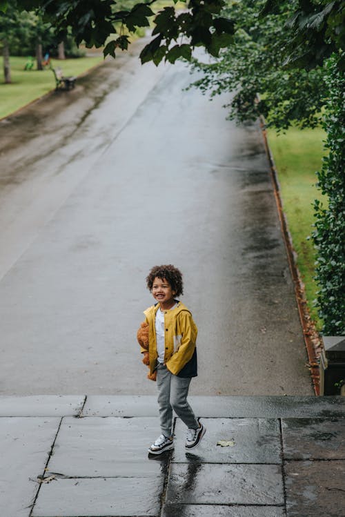 Cheerful ethnic boy walking on wet pavement in city park