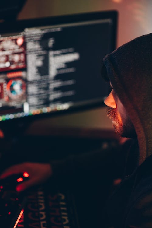 Free Side view of crop anonymous cyber spy in hoodie hacking computer system with information on screen at night Stock Photo