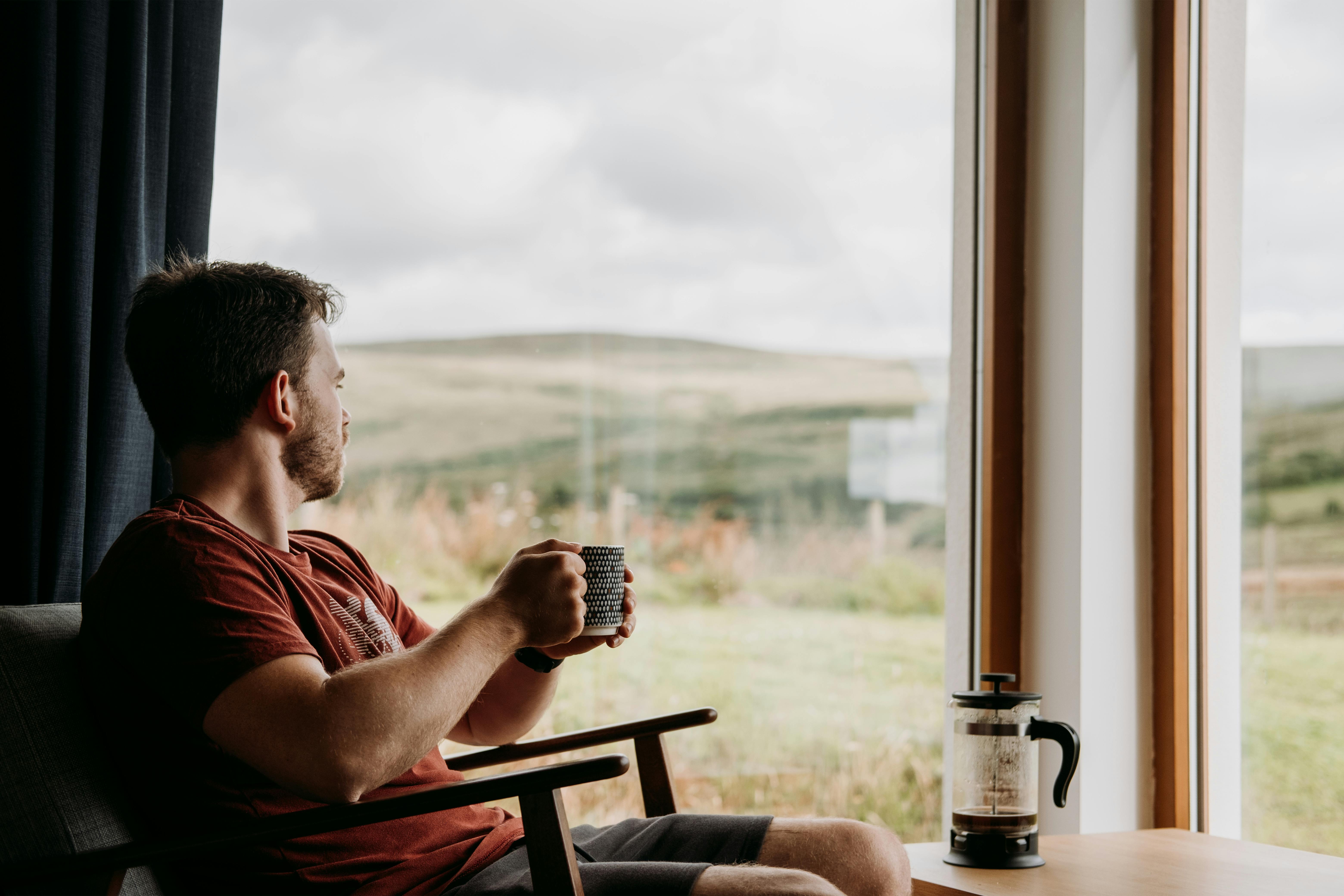 Side view of young dreamy male with mug of hot beverage looking away against glass wall in countryside house by Anete Lusina