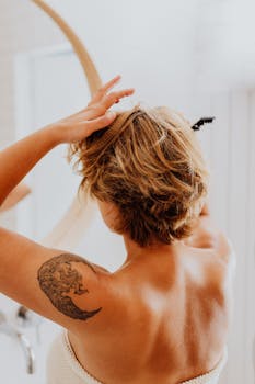 Back view of a woman with a tattoo adjusting her hair in a bright bathroom mirror.