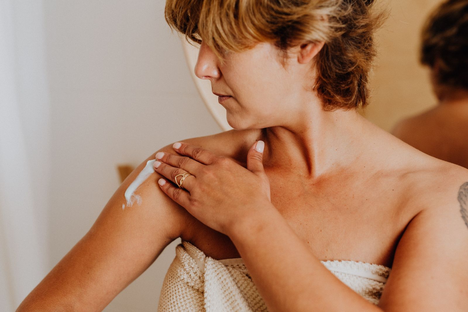 Woman with short blonde hair applying skincare cream to her shoulder in warm natural light.