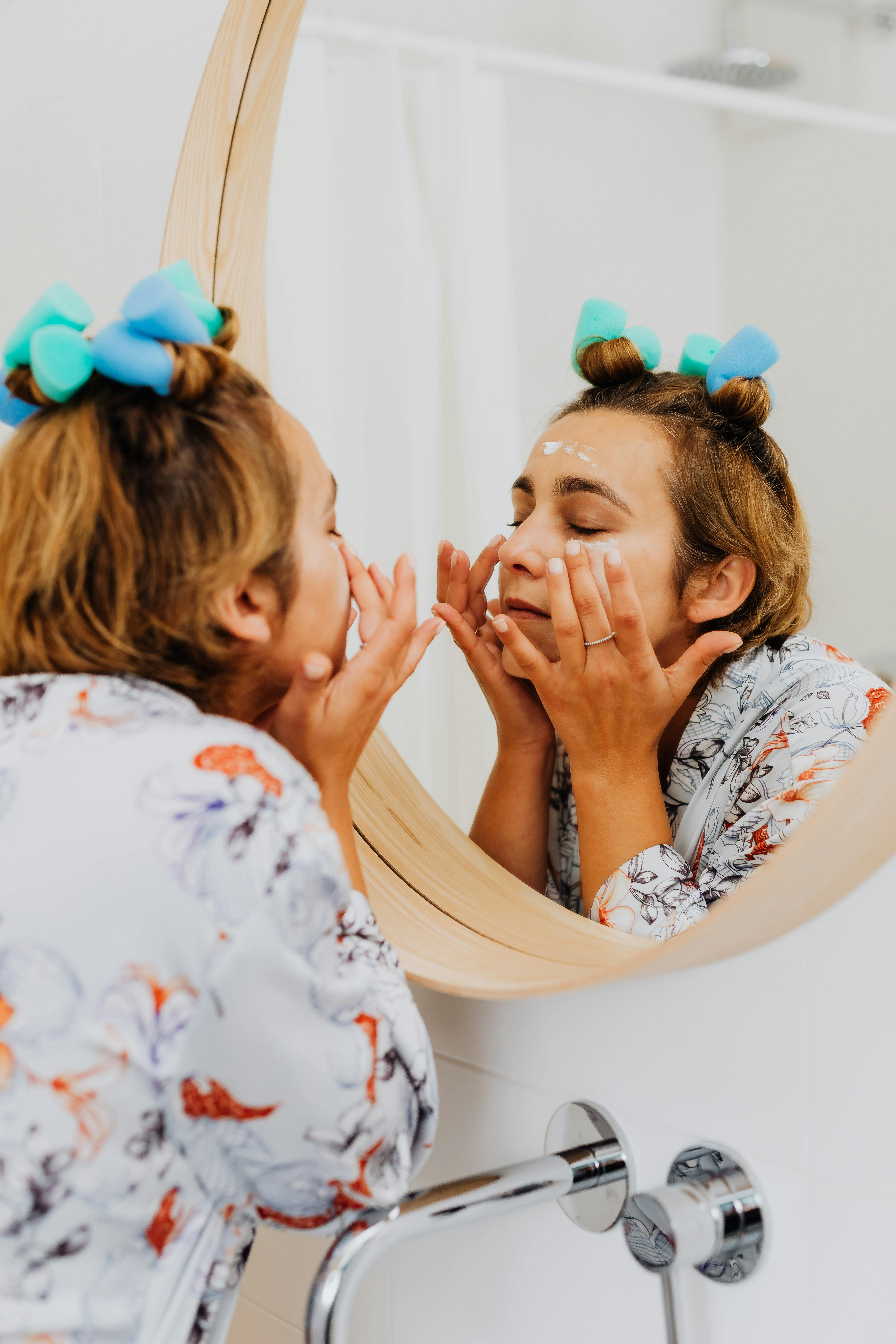 Woman in a robe applies face cream with curlers in hair, focusing on skincare routine.