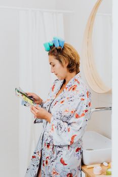 A woman in a floral robe with hair rollers reading a magazine indoors.