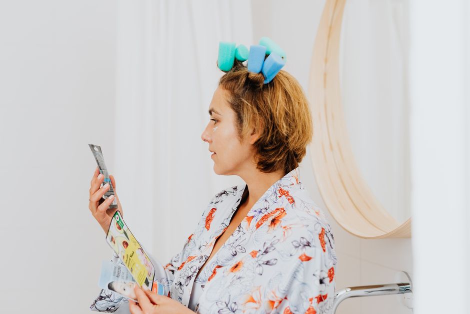 Woman in a bathrobe with hair curlers reads skincare instructions indoors.