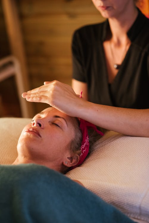 Free Relaxed woman with closed eyes lying under blanket while getting reiki therapy treatment from unrecognizable therapist in cozy salon with blurred background Stock Photo