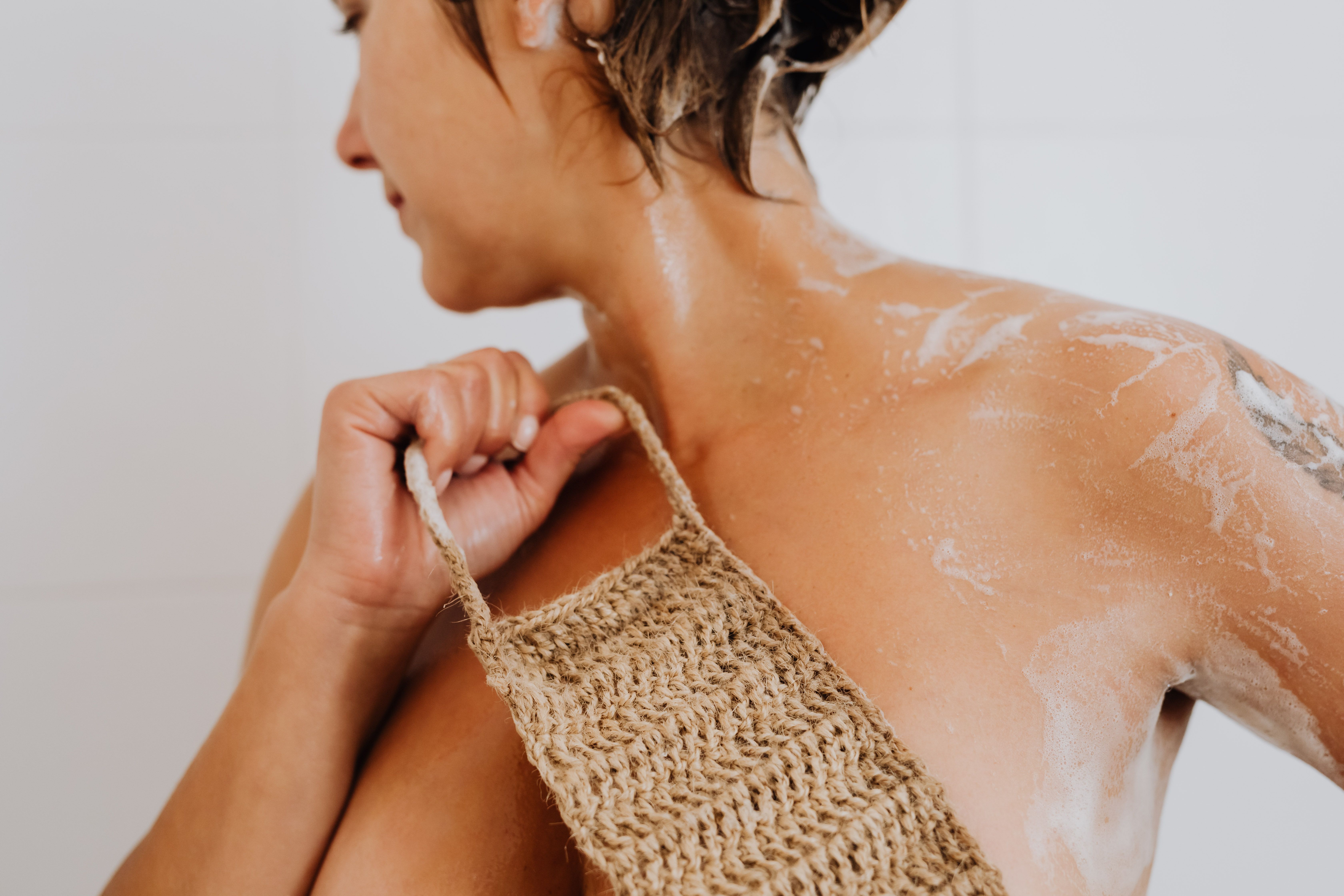 Adult woman with short hair exfoliates her skin using a loofah in the shower.
