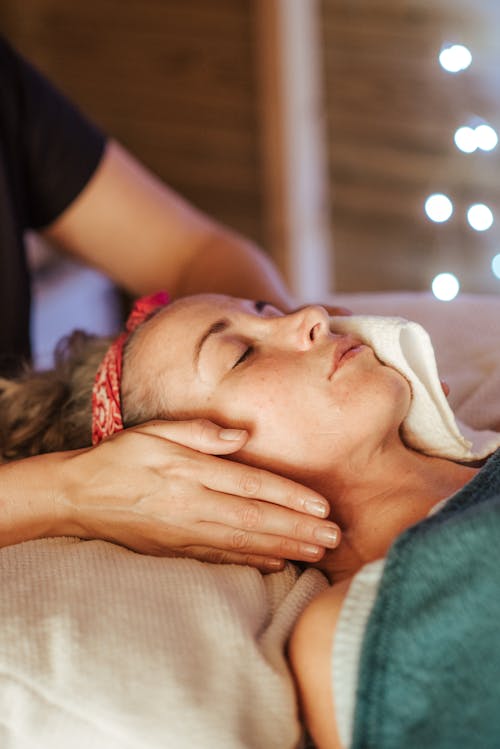 Free Adult female resting on comfortable bed in spa salon while having face massage from masseur Stock Photo