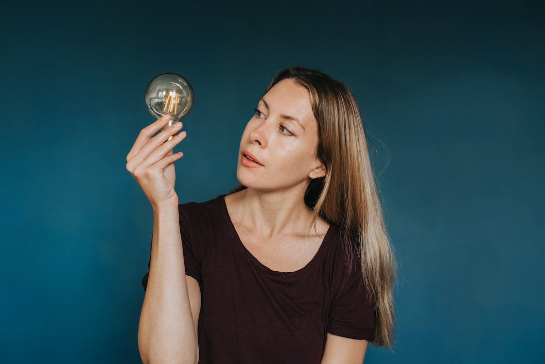 Free Enthusiastic female in casual clothes standing against blue background and looking at glowing light bulb Stock Photo