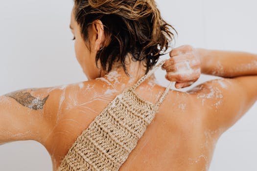 Back view of a woman with tattoos exfoliating her skin with a loofah in the shower.