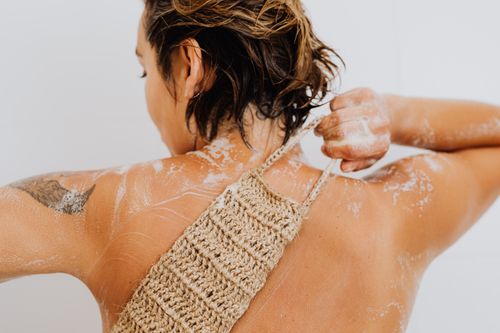 Free Back view of a woman with tattoos exfoliating her skin with a loofah in the shower. Stock Photo