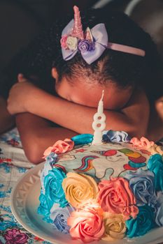 一个面目全非的非裔美国小女孩，用手臂上的头巾遮住脸，坐在桌子旁，手捧生日蛋糕许愿