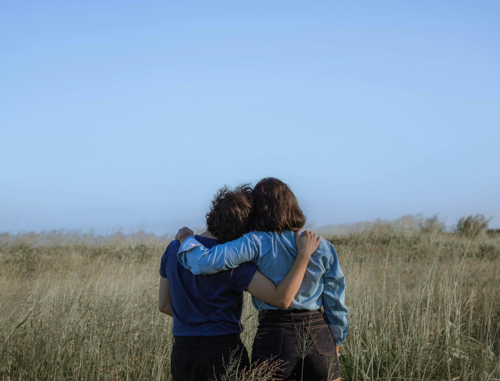 Back view unrecognizable girlfriends wearing casual clothes hugging each other while standing on grassy vast field in summer countryside