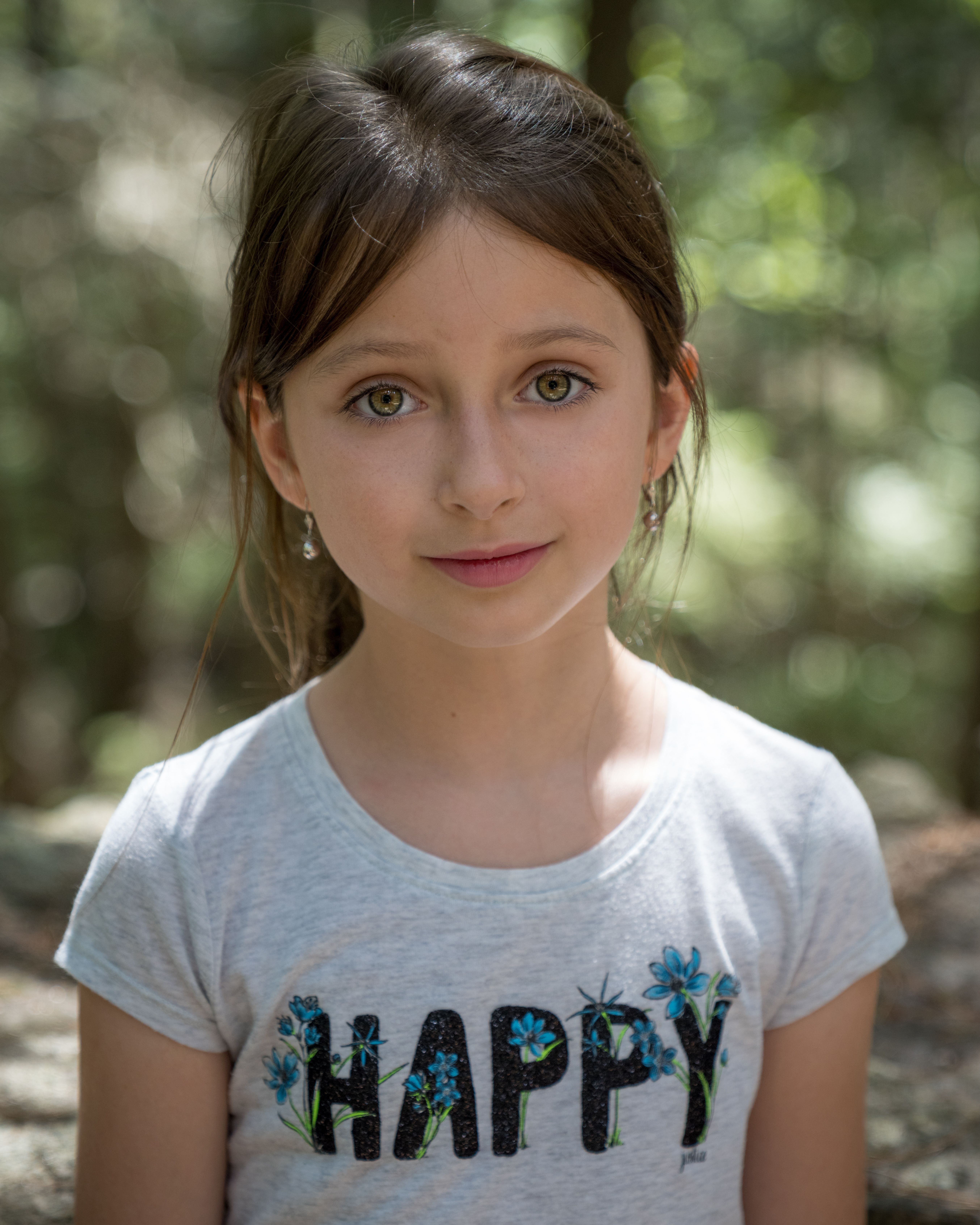 Emotionless girl in casual t shirt in park near green trees in summer sunny day looking at camera