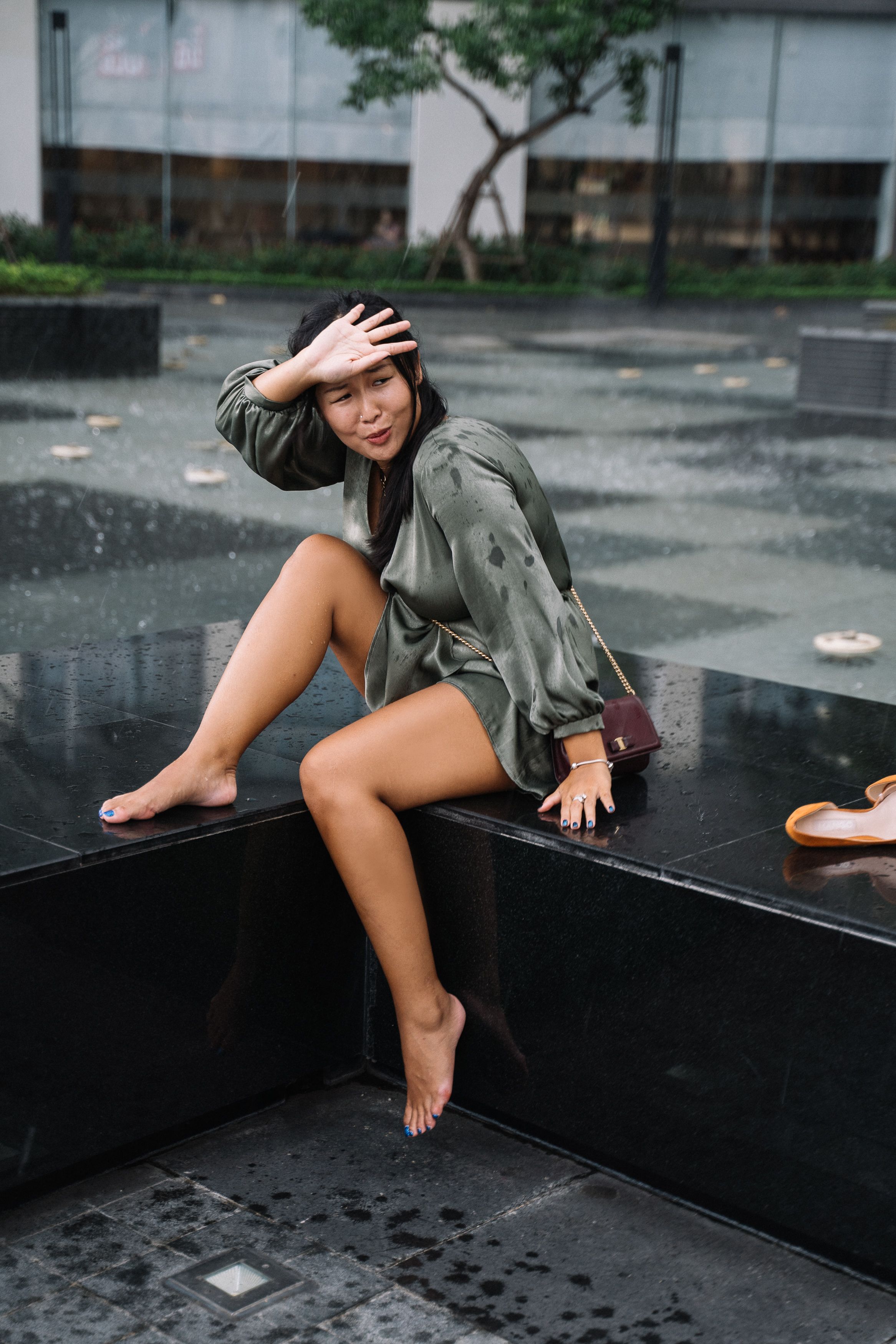 一名年轻女子身穿绿色连衣裙，赤脚坐在城市喷泉旁，享受着雨水
