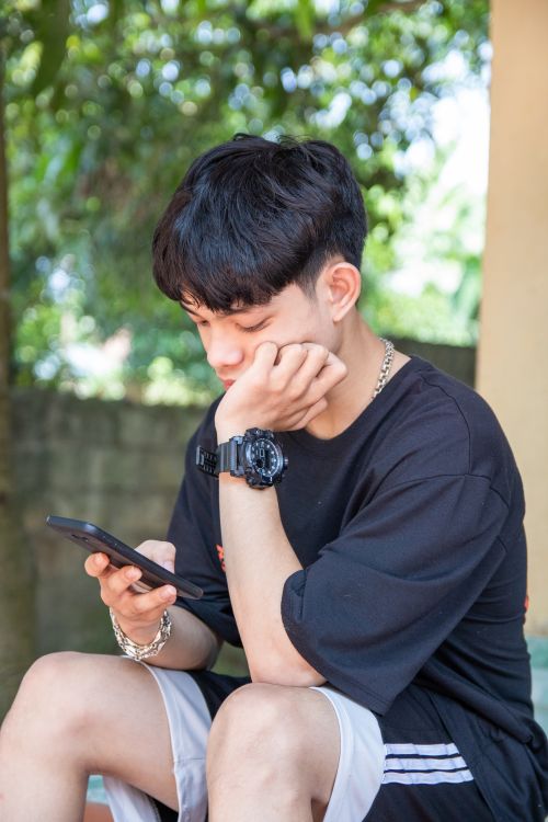 Free A young male teenager engrossed in his smartphone while sitting outdoors in a casual setting. Stock Photo