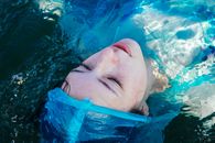 From above of young dead female with makeup and closed eyes in blue raincoat lying in river with bubbles on surface