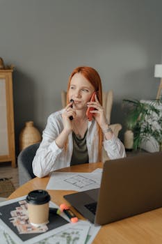 Woman multitasking with smartphone and laptop during remote work in home office setting.