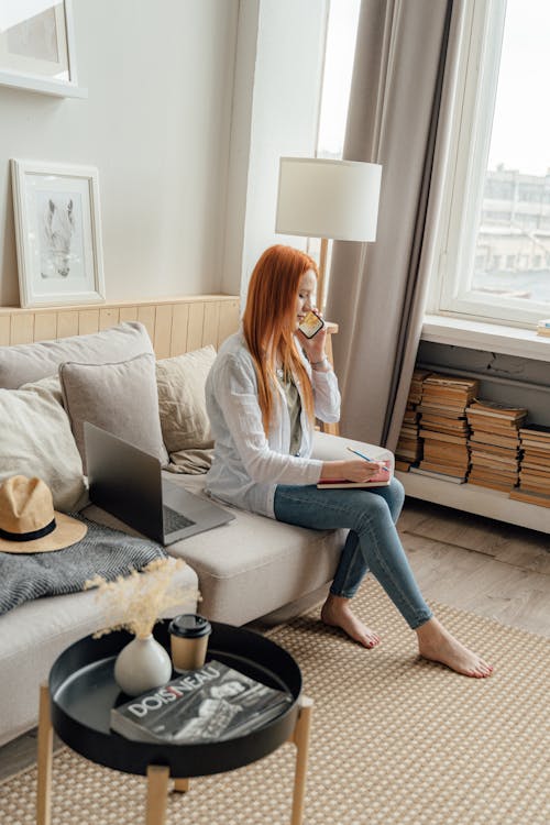 Woman in White Long Sleeve Shirt and Blue Denim Jeans Sitting on Gray Couch Talking on Phone 