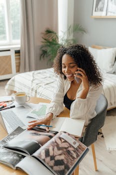 Young woman juggling phone call, magazine browsing, and work on laptop at home office.