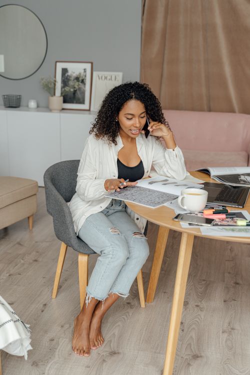 Free African American woman multitasking with a laptop and phone call at home, smiling and seated at a modern table. Stock Photo