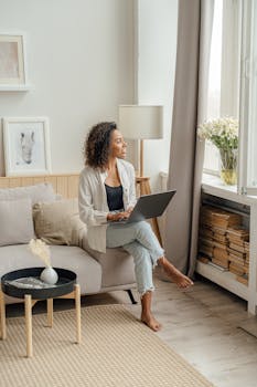 African American woman working on laptop at home, enjoying natural light by the window.