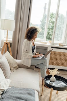 A woman sitting comfortably on a sofa using a laptop in a bright, cozy living room.