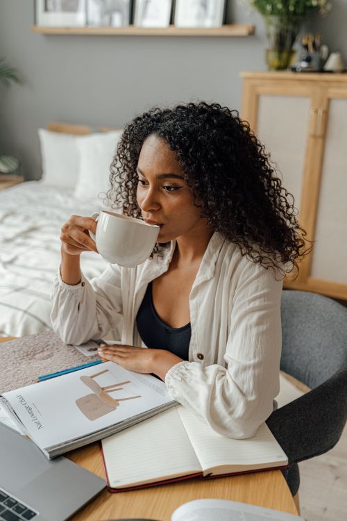 Free Woman drinking coffee and working on her laptop with books spread out on a cozy desk at home. Stock Photo