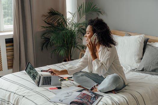 A woman works from home, multitasking on her laptop and phone while sitting on her bed.
