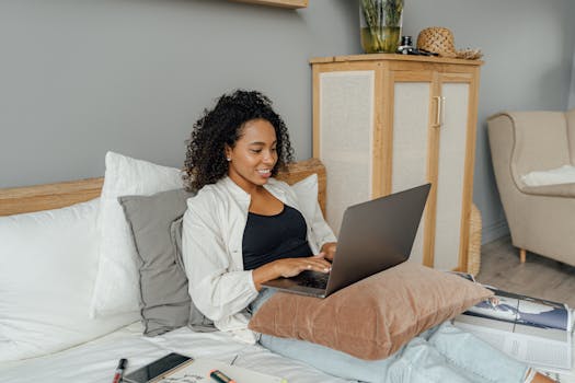 A young woman sits on her bed working remotely using a laptop, creating a cozy home office space.