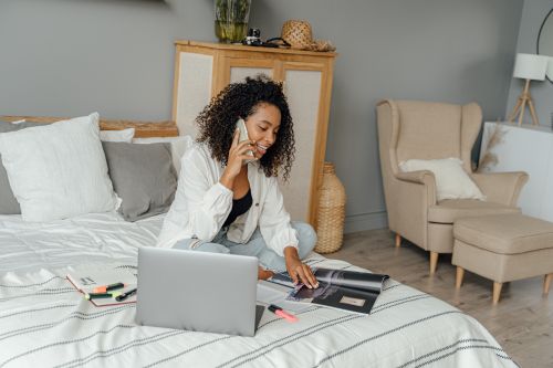 Free Black woman multitasking with laptop and phone, working remotely in a chic modern bedroom. Stock Photo