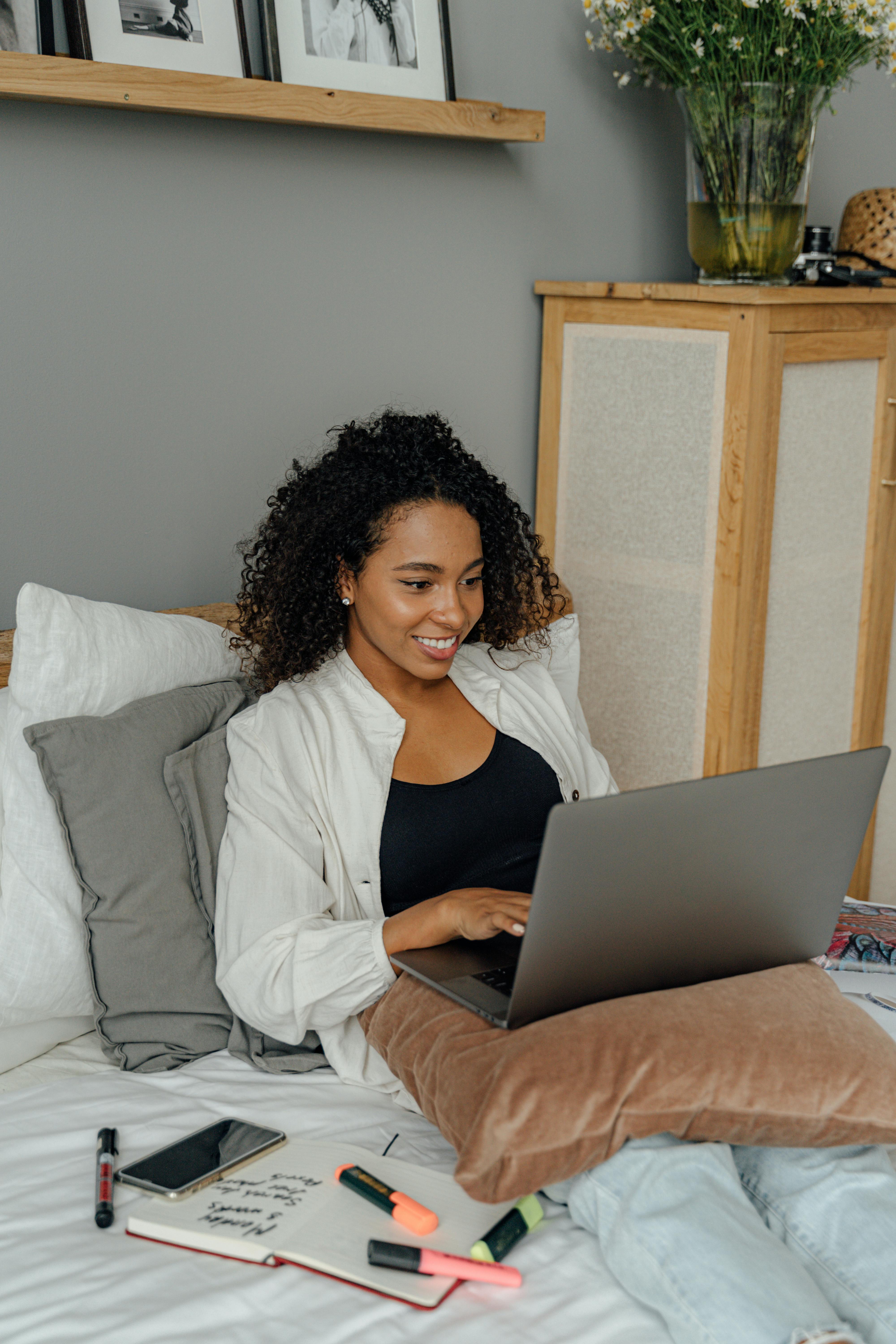 African American woman relaxing and working on a laptop in a cozy bedroom setting.