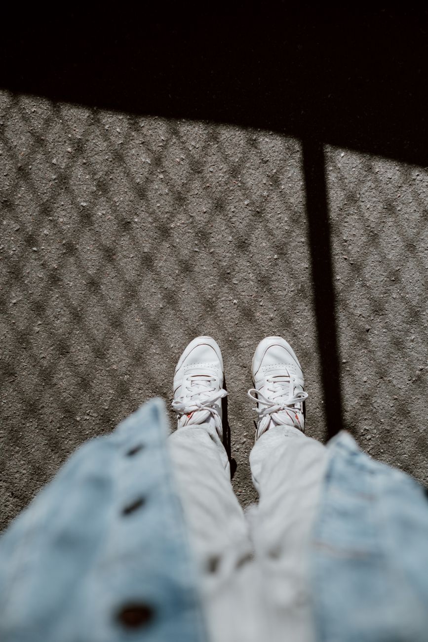 A top-down view of stylish white sneakers on a sunlit pathway with shadow patterns.