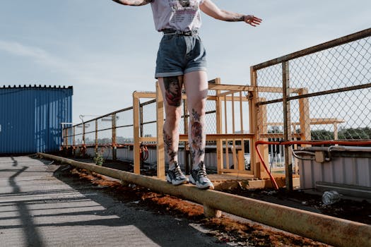 A person with tattooed legs balances on a pipeline in an industrial outdoor setting.