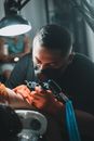 Crop serious male master in protective mask wearing gloves using special machine to make tattoo on arm of client lying under lamp with blurred background