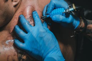 High angle hands of anonymous tattooist in blue gloves using professional tool to make tattoo on back of client in salon