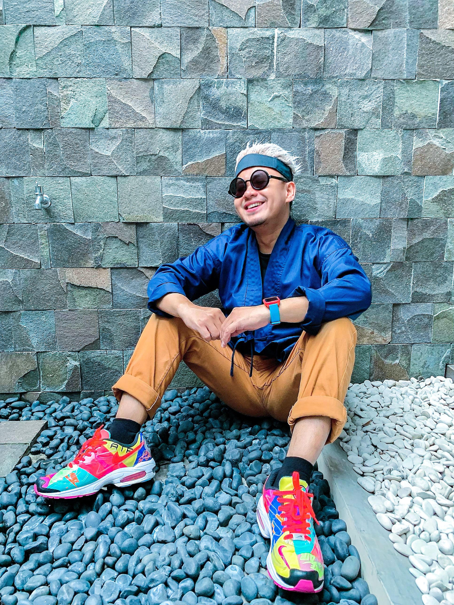 Smiling man in colorful sneakers and bandana sitting on stones against a stone wall.