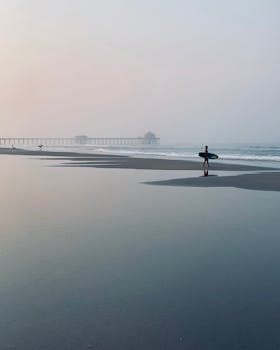A lone surfer carrying a surfboard on Huntington Beach at sunset with the iconic pier in the background.